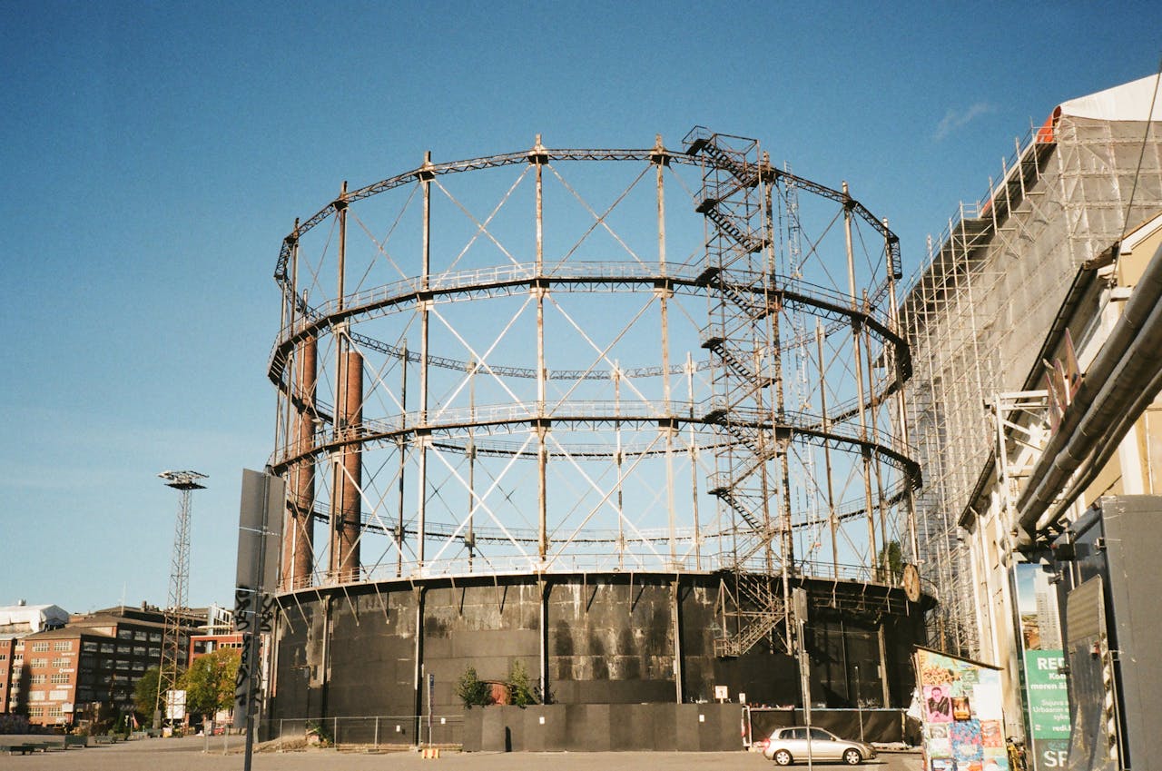 A large industrial gasometer structure in an urban setting under a clear blue sky.